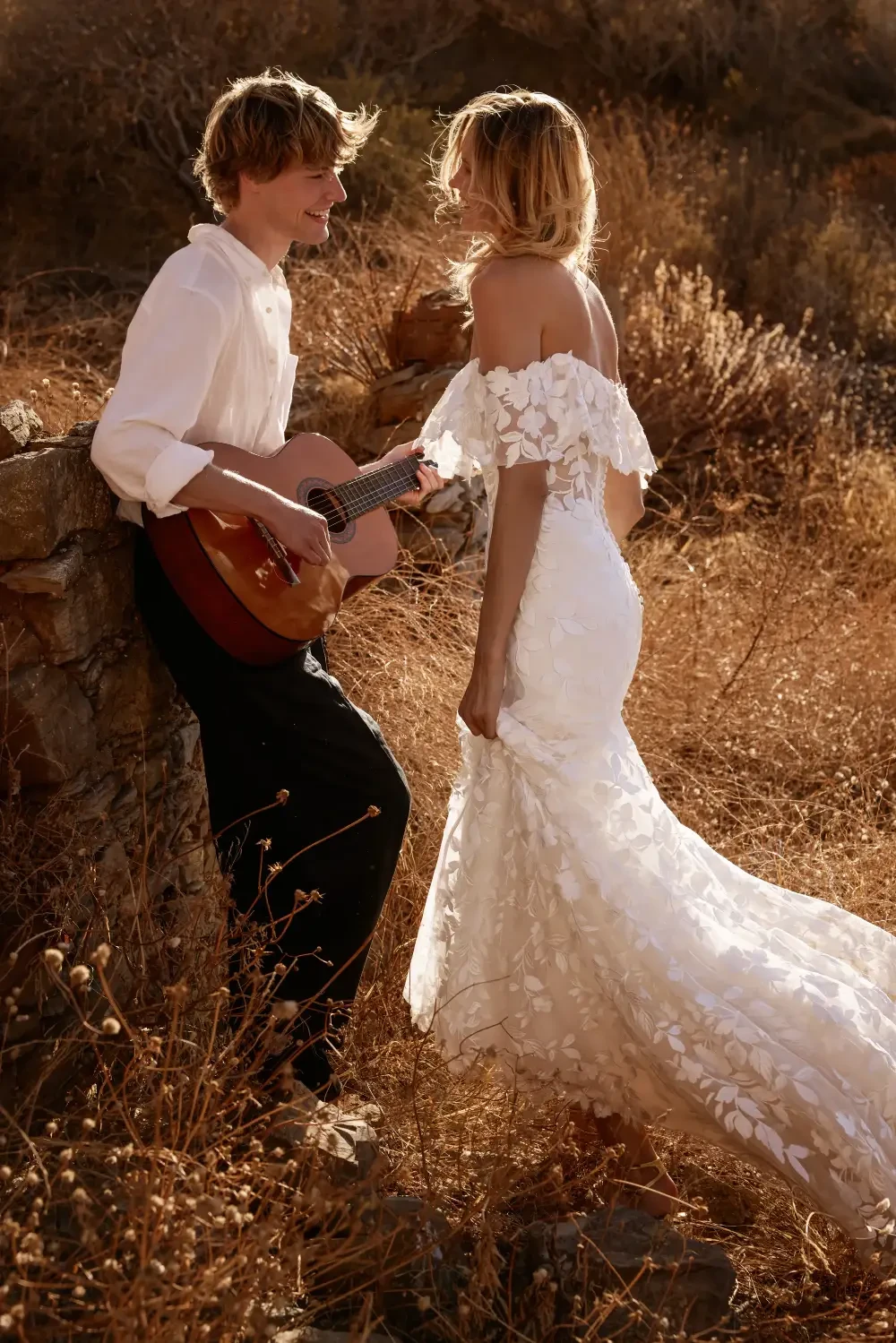 A couple in a romantic pose outdoors, with the man playing a guitar and the woman wearing a flowing white dress. The setting has dry grass and a rocky backdrop.