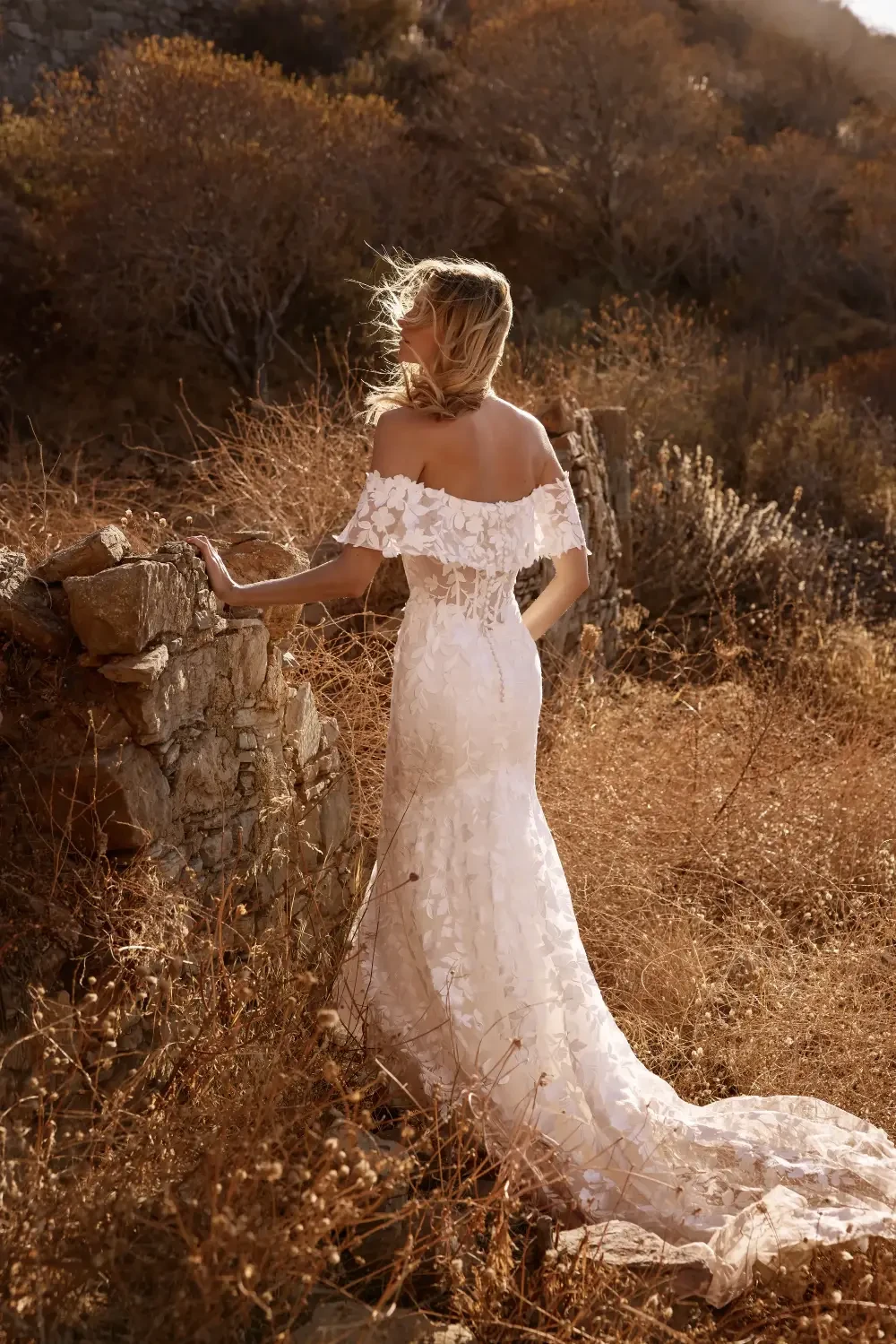 A woman in a flowing white dress standing near a stone wall in a dry, grassy landscape with autumn foliage.