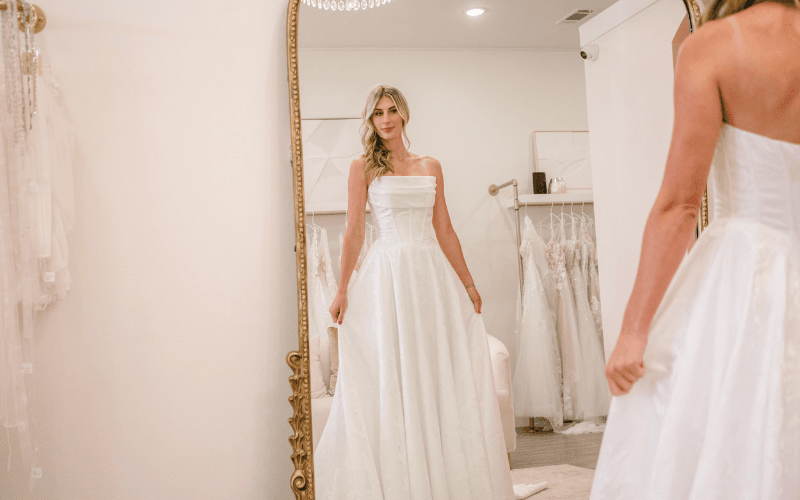 Bride trying on a strapless satin A-line wedding dress with a pleated bodice and full skirt, standing in front of a mirror in a bridal boutique.