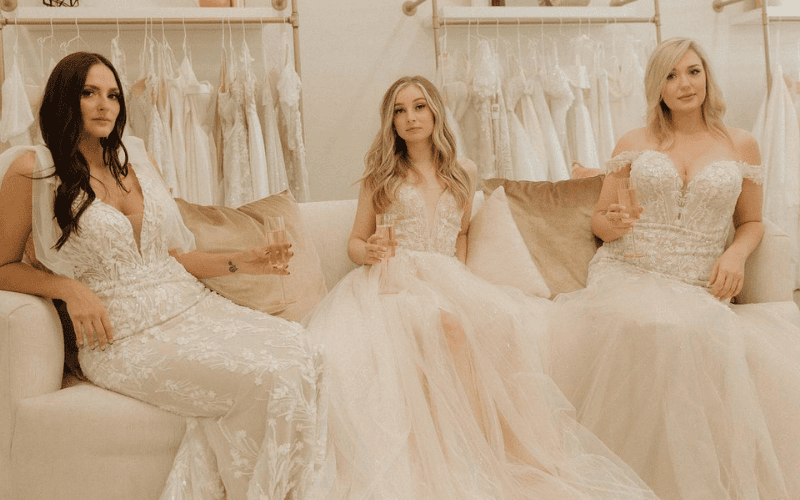 Three brides wearing lace wedding dresses sit elegantly on a sofa, each holding a glass of champagne, with a backdrop of hanging gowns in a bridal boutique.