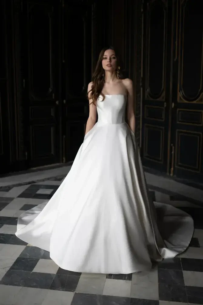 A woman in a strapless white gown standing in an elegant, dimly lit room with dark wood paneling and a patterned floor.