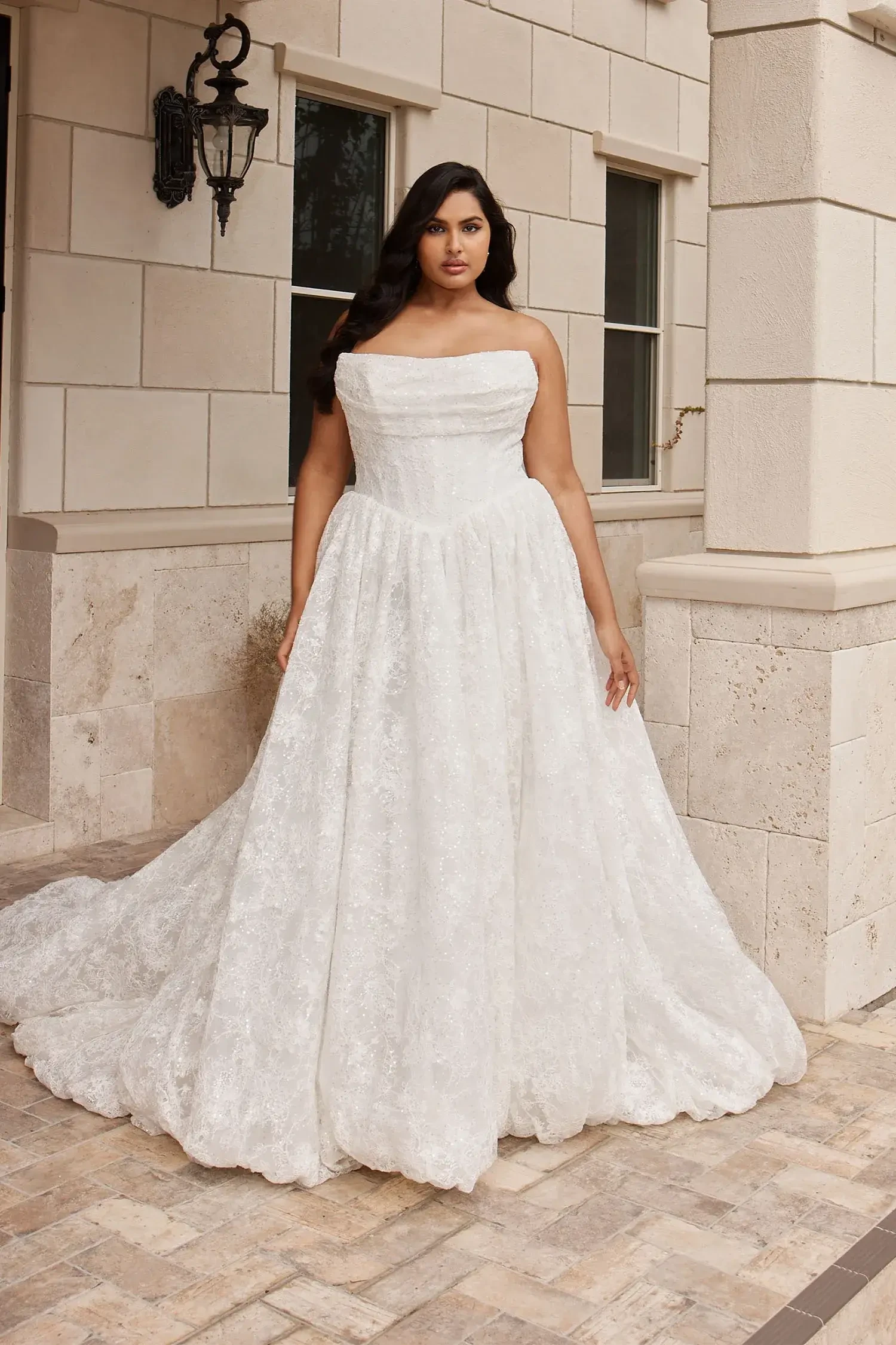 A person wearing a white strapless gown stands outside near a building with light-colored stone walls. The gown has a flowing skirt and intricate details, and the person appears confident in their pose.