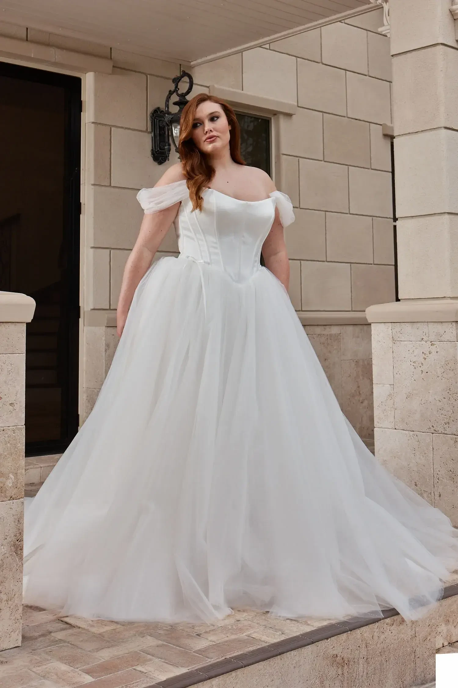 A woman in a flowing white gown stands on a staircase outside a building. The gown features off-the-shoulder sleeves and has a fitted bodice with a voluminous skirt.