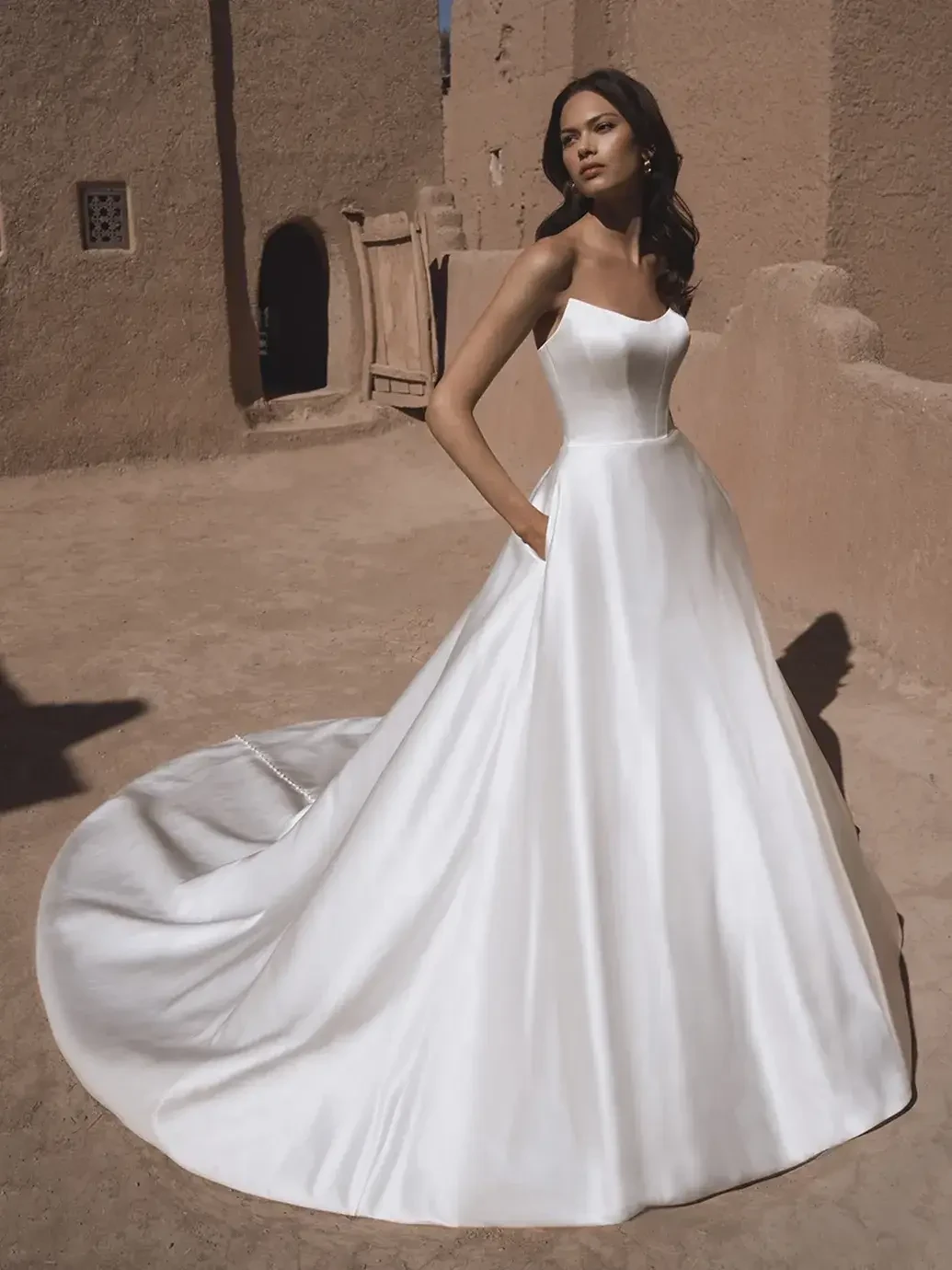 A model wearing a sleek, strapless white wedding gown with pockets, standing in an outdoor setting with earthy tones and rustic architecture.