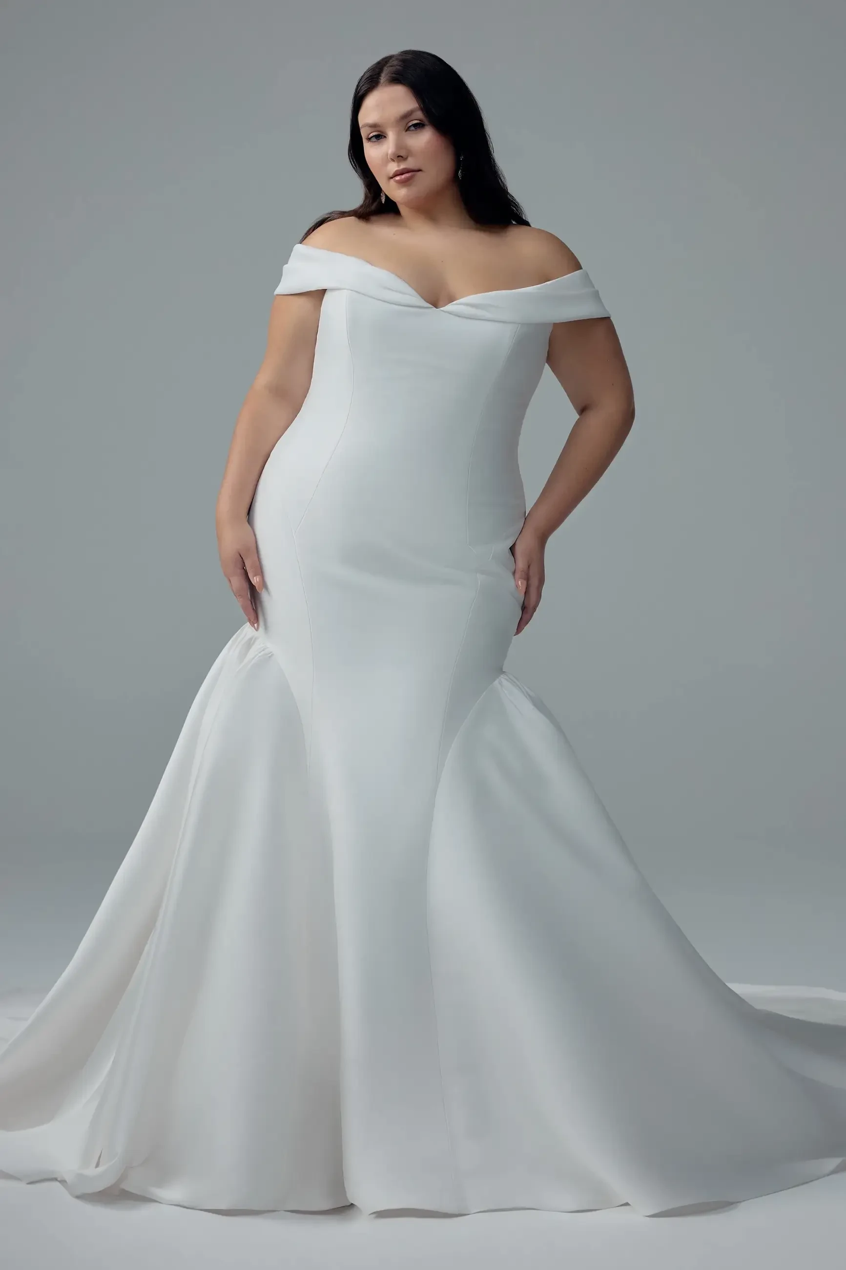 A woman wearing an off-the-shoulder white gown, standing confidently with a neutral background.